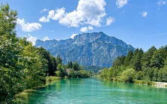 Turquoise Königsseeache river near Anif with rocky mountains in the background, framed by green trees under a blue sky with white clouds.