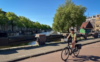 Female cyclist riding across a bridge over an Amsterdam canal. Tree-lined canal and traditional Dutch buildings visible in the background.