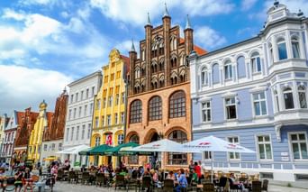 Bunte historische Gebäude mit gotischer Backsteinarchitektur säumen einen Marktplatz in Stralsund. Straßencafés mit Sonnenschirmen und Besucher füllen den Platz.