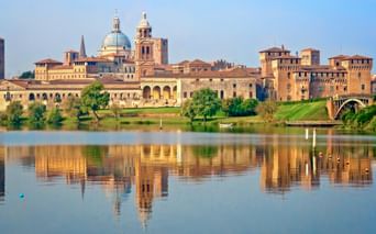 Historische Stadtansicht von Mantua mit Domkuppel und Glockenturm, die sich im ruhigen Seewasser spiegeln. Grüne Rasenflächen und Bäume säumen das Ufer.
