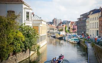 Canal in Ghent with colorful buildings, moored boats, and tourists on a boat tour. Trees line the waterfront under a blue sky with clouds.