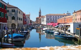 Kanal in Chioggia mit vertäuten Booten und bunten Gebäuden. Ein Kirchturm erhebt sich im Hintergrund unter blauem Himmel.