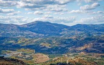 Weitläufige Berglandschaft bei Zaros, Kreta, mit sanften Hügeln voller Olivenhaine und landwirtschaftlichen Feldern unter blauem Wolkenhimmel.