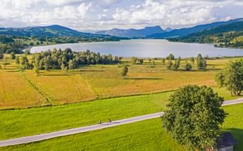 Two cyclists on a country road through green meadows with Irrsee lake and mountains under cloudy sky in Austrian Seenland region.