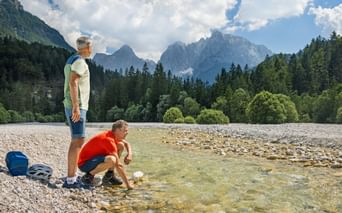 Two cyclists resting by a clear mountain river with pebble beach. Dense forest and dramatic mountain peaks visible under cloudy sky.