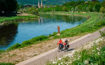 Zwei Radfahrer auf Weg entlang der Weser mit Angler am Flussufer. Grüne Hügel und Kirchtürme von Höxter im Hintergrund sichtbar.