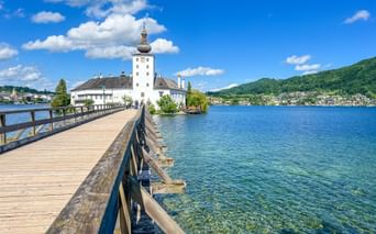 Holzsteg führt zu Schloss Orth am Traunsee im Salzkammergut. Klares blaues Wasser, weißes Schloss mit Turm, grüne Hügel.