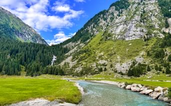 Turquoise Krimml Ache river flowing through green alpine meadow with forested mountains and rocky peaks under blue sky.
