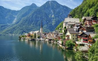 Hallstatt mit traditionellen Häusern und Kirchturm am Ufer eines türkisfarbenen Sees, umgeben von grünen bewaldeten Bergen.