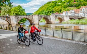 Zwei Radfahrer fahren entlang der Weser in Hann. Münden mit der historischen Werrabrücke und Fachwerkhäusern am Hang.