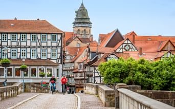 Zwei Radfahrer überqueren eine Steinbrücke zur Altstadt von Hann. Münden mit Fachwerkhäusern und Kirchturm unter klarem blauen Himmel.