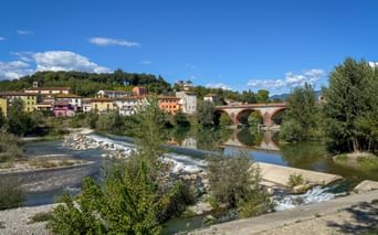 Stone bridge with arches spanning a river in Tuscany. Colorful buildings line the hillside behind the bridge, with trees and rocky riverbed in foreground.