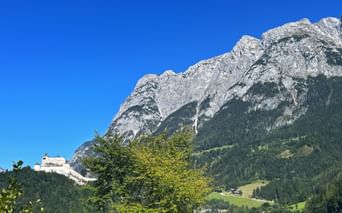 Burg Werfen auf einem bewaldeten Hügel mit den dramatischen Felsgipfeln des Tennengebirges dahinter unter klarem blauen Himmel.