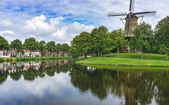 Traditional Dutch windmill Molen de Hoop on green lawn beside calm canal reflecting trees and houses in Middelburg under blue sky.