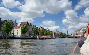 View from a boat on Amsterdam's Amstel River showing historic canal houses, tour boats, and blue sky with white clouds.