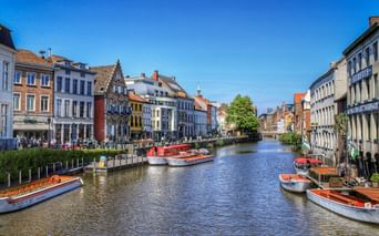 Kanal in Gent gesäumt von bunten historischen Gebäuden und Ausflugsbooten. Menschen sitzen am Ufer unter blauem Himmel.