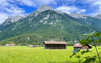 Green alpine meadow with traditional wooden barn in Bavaria. Mountains with rocky peaks and forests rise behind scattered farm buildings under blue sky.