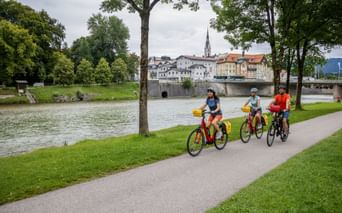 Drei Radfahrer fahren entlang der Isar in Bad Tölz. Historische Stadt mit Kirchturm und Brücke über dem Wasser, eingerahmt von grünen Bäumen.
