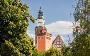 Roter Turm mit weißem Oberteil und dunkler Spitze neben Fachwerkkirche in Kamenz, eingerahmt von grünen Bäumen unter blauem Himmel.