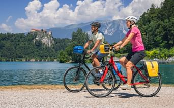 Two cyclists with panniers riding along Lake Bled shore. Bled Castle on cliff and mountains visible in background under blue sky with clouds.