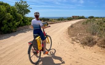 Cyclist with helmet and yellow panniers on sandy coastal path in Alto Minho, Portugal. Ocean visible in background under blue sky.