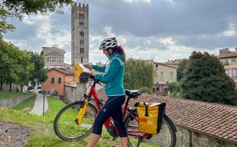 Female cyclist with yellow panniers on red bike in Lucca, Tuscany. Behind her rises a tall medieval bell tower against a cloudy sky.