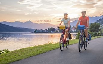 Two cyclists riding along lakeside path at Millstätter See during sunset, with mountains and village in background.