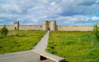 Wooden boardwalk leading through green marshland to the fortified walls of Aigues-Mortes with towers and gates under a cloudy sky.