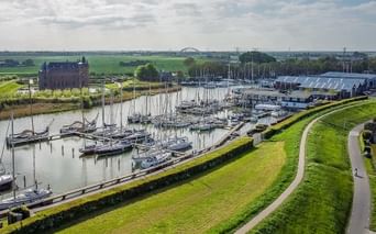 Luftaufnahme des Hafens von Muiden mit Segelbooten und Yachten entlang grüner Deiche. Historisches Schloss Muiderslot links sichtbar.