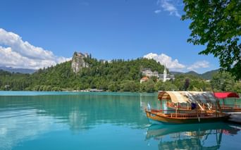 Traditional wooden boat on turquoise Lake Bled in Slovenia with Bled Castle on forested cliff and white church spire visible on shore.