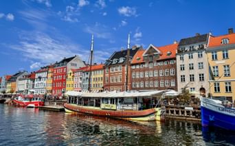 Historic Nyhavn harbor in Copenhagen with colorful townhouses, boats moored along the canal, and outdoor cafes under white awnings on a sunny day.