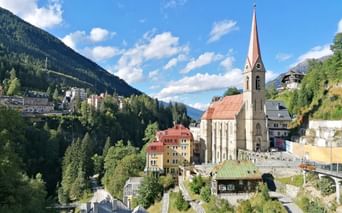 Church with distinctive red spire in Bad Gastein, surrounded by colorful buildings on a hillside with forested mountains and blue sky.