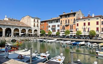 Harbor of Desenzano at Lake Garda with moored boats, colorful historic buildings, and white arched colonnade under clear blue sky.