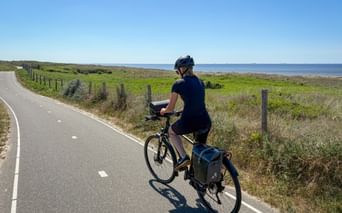 Female cyclist with panniers riding on paved coastal bike path in Zeeland, with green meadows, fence posts, and blue sea in background.