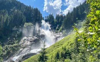 Upper Krimml Waterfalls cascading down rocky slopes surrounded by green meadows and dense forests under a blue sky with white clouds.