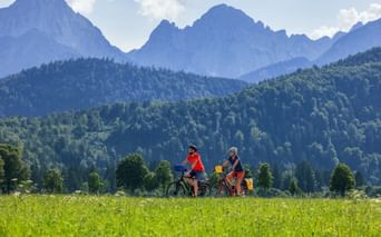 Two cyclists riding through a green meadow with forested hills and dramatic mountain peaks of the Salzburg Alps in the background under a blue sky.