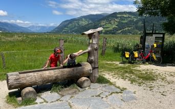 Cyclist in red with child at rustic wooden fountain in Schüttdorfer Moor. Touring bike with yellow panniers parked nearby, mountains in background.