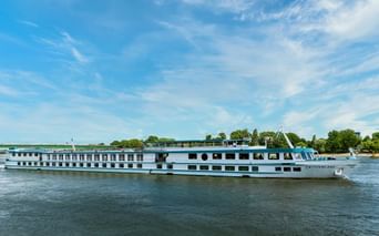 White river cruise ship MS Switzerland sailing on calm water with blue sky. The multi-deck vessel features rows of windows and a sun deck.