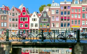 Reihe bunter historischer Grachtenhäuser in Amsterdam, die sich im Wasser spiegeln. Fahrräder stehen am Brückengeländer im Vordergrund.