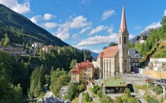 Church with tall spire in Bad Gastein surrounded by colorful buildings on hillside. Green mountains and blue sky with clouds frame the alpine town.