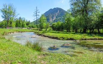 Clear river Ammer flowing through green meadows with trees and a forested mountain peak under blue sky near Munich, Bavaria.