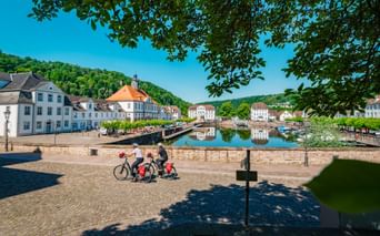 Three cyclists at the baroque harbor in Bad Karlshafen with historic white buildings, a canal basin, and green hills under blue sky.