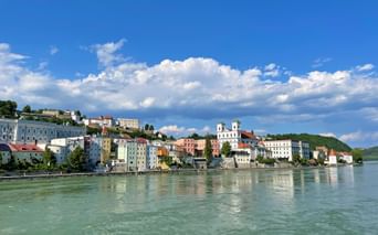 Bunte Gebäude von Passau entlang der Donau mit sichtbarer weißer Kirche und Türmen. Blauer Himmel mit weißen Wolken über der Hangstadt.