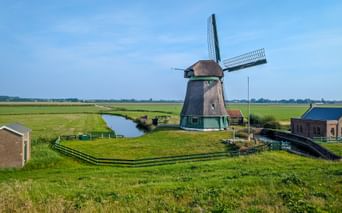 Dutch windmill in Schellinkhout surrounded by green polder landscape with canal and farm buildings under blue sky in North Holland.