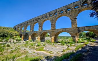 The ancient Roman Pont du Gard aqueduct with three tiers of arches spanning across a dry riverbed in Provence under a clear blue sky.