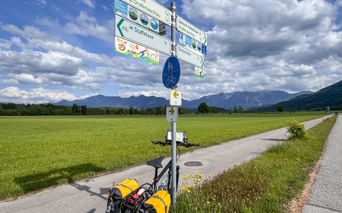 Bicycle with yellow panniers at directional signpost in Murnauer Moos. Green fields and mountains under cloudy sky in background.
