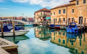 Bunte Boote am ruhigen Kanal in Murano mit traditionellen Backsteingebäuden, die sich im Wasser spiegeln, unter blauem Himmel.