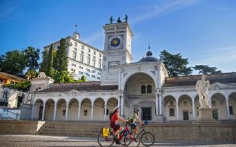 Two cyclists riding past the historic clock tower and colonnade at Piazza della Libertà in Udine, Friuli, under a blue sky.
