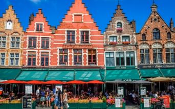 Row of traditional Flemish gabled houses with red, beige, and brown facades at Bruges Grote Markt. Green awnings shade outdoor cafes with red umbrellas.