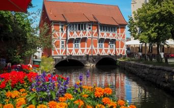 Orange half-timbered house with red tile roof spanning a canal in Wismar. Colorful flowers bloom in the foreground along the waterway.